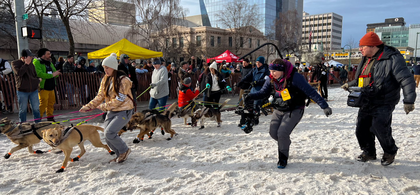 FrostlineStudios_Iditarod-2025_03-03-2025_2 Frostline Studios crew run alongside a dog team at the Iditarod ceremonial start in Anchorage, Alaska.